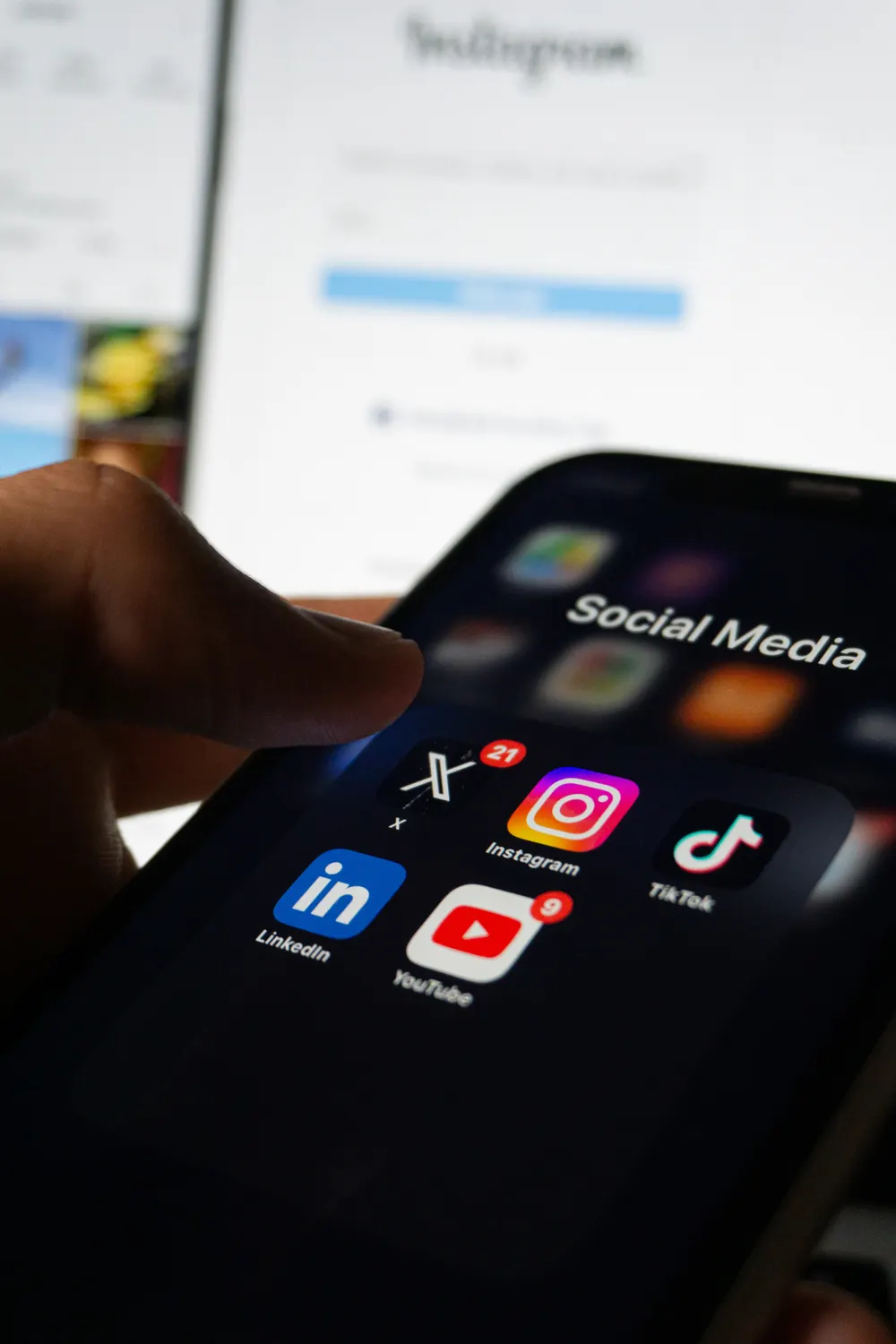 A hand reaching for a phone with a Social Media folder of X, Instagram, TikTok, LinkedIn and YouTube